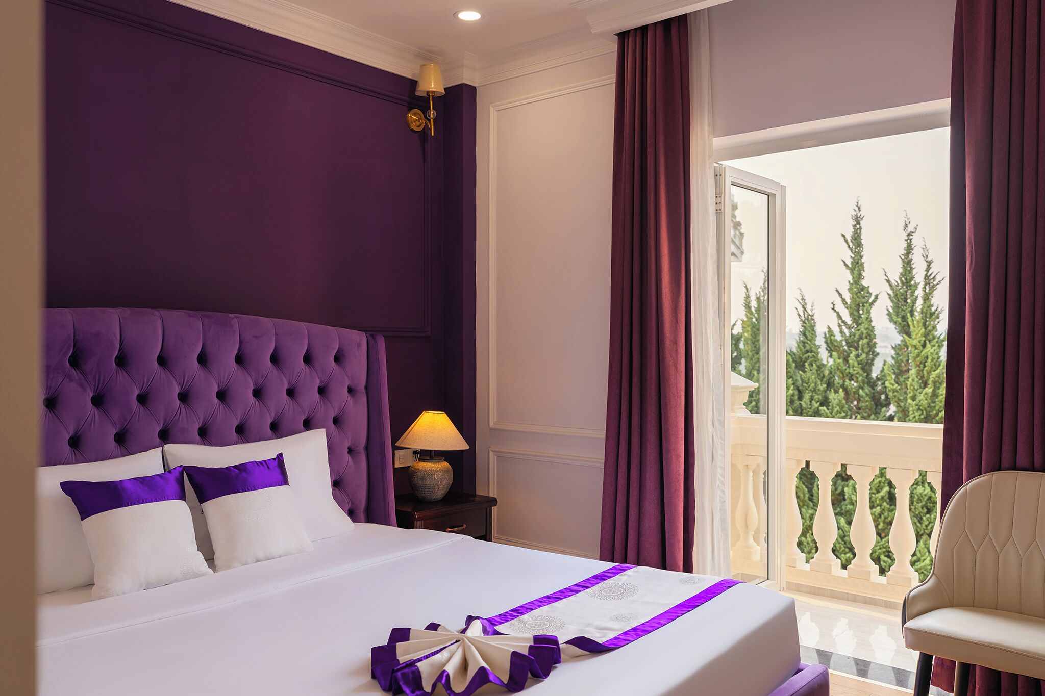 A hotel room with a large bed, a nightstand with a lamp, a balcony with a railing, and a view of trees.