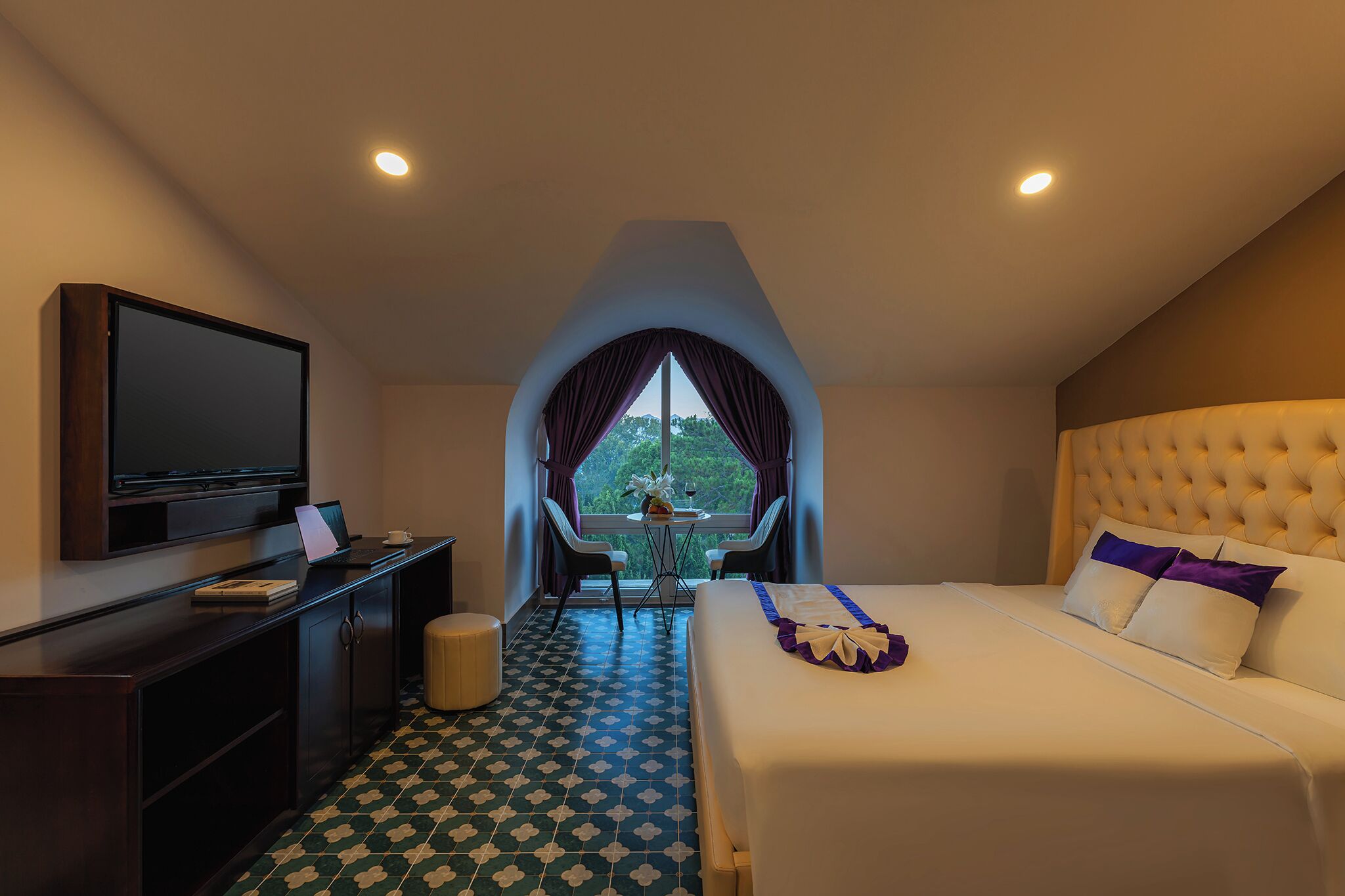 A hotel room with a bed, a TV, a desk with a laptop, and a view of trees through a window.