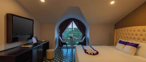 Habitación de hotel con cama, televisión, escritorio con computadora portátil y vista a árboles por la ventana.