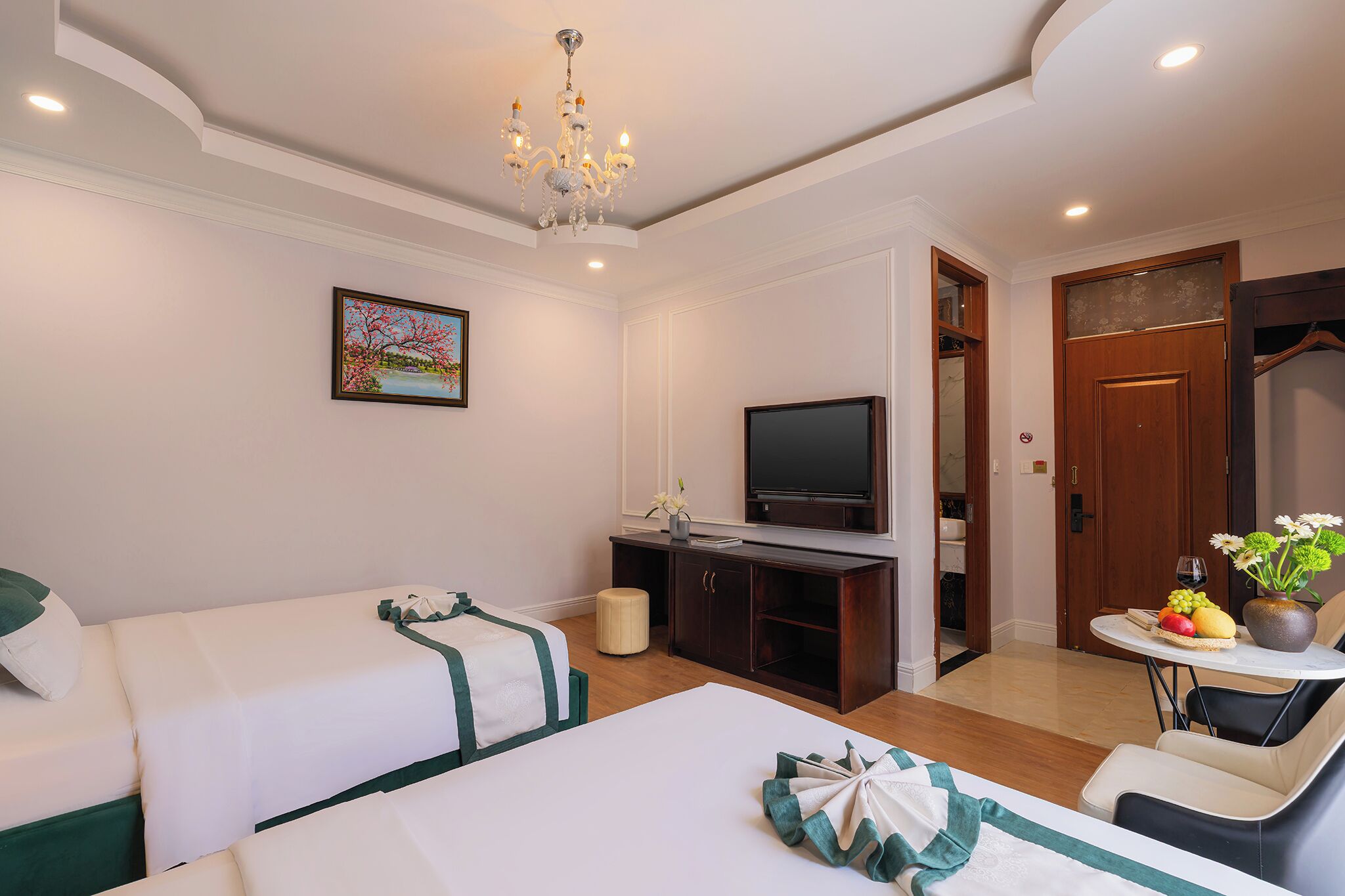 A hotel room with two beds, a television, a dining table with a fruit bowl, and a chandelier.