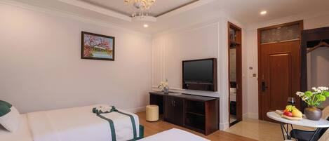 A hotel room with two beds, a television, a dining table with a fruit bowl, and a chandelier.