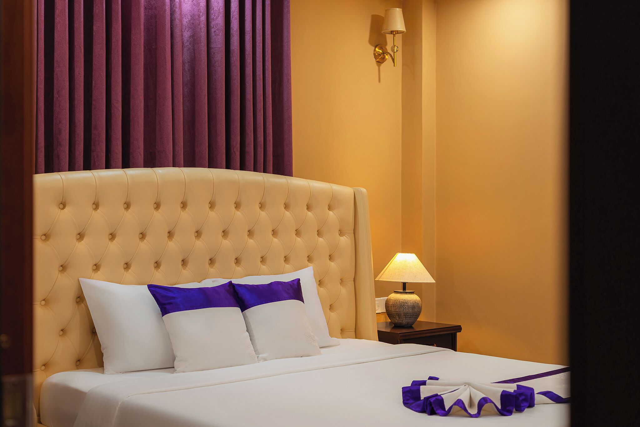 A bed with a tufted headboard, white bedding, and purple accents, a nightstand with a lamp, and a wall-mounted light fixture.