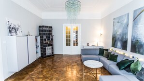 Un vaste espace de vie doté d’un parquet à motifs en chevrons, d’un canapé gris agrémenté de coussins verts, d’une table basse blanche et d’un lustre.