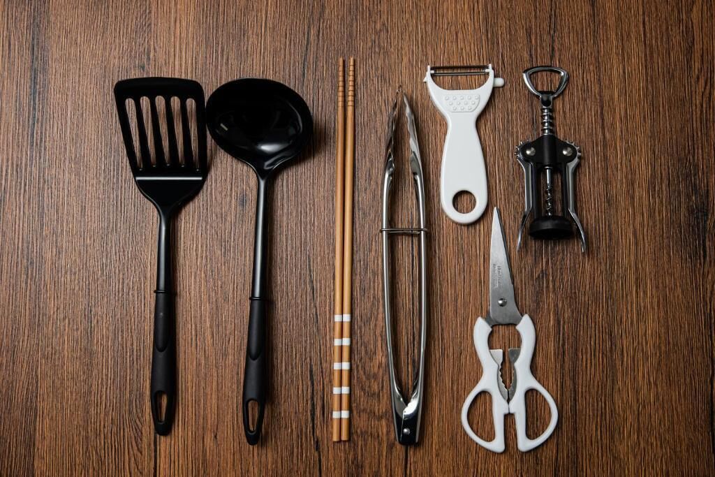 A wooden surface with kitchen utensils including a black spatula, a black ladle, a pair of chopsticks, a metal grater, a white grater, a pair of scissors, and a black corkscrew.