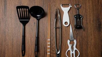 A wooden surface with kitchen utensils including a black spatula, a black ladle, a pair of chopsticks, a metal grater, a white grater, a pair of scissors, and a black corkscrew.