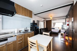 A compact room with a kitchenette, a dining table, a bed, and a bedside table with a lamp. - Vacation Rental NISHIDA (Kagoshima)