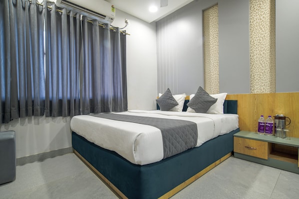 A neatly made bed with a blue headboard, a bedside table with bottles and a thermos, and a grey curtain covering a window.