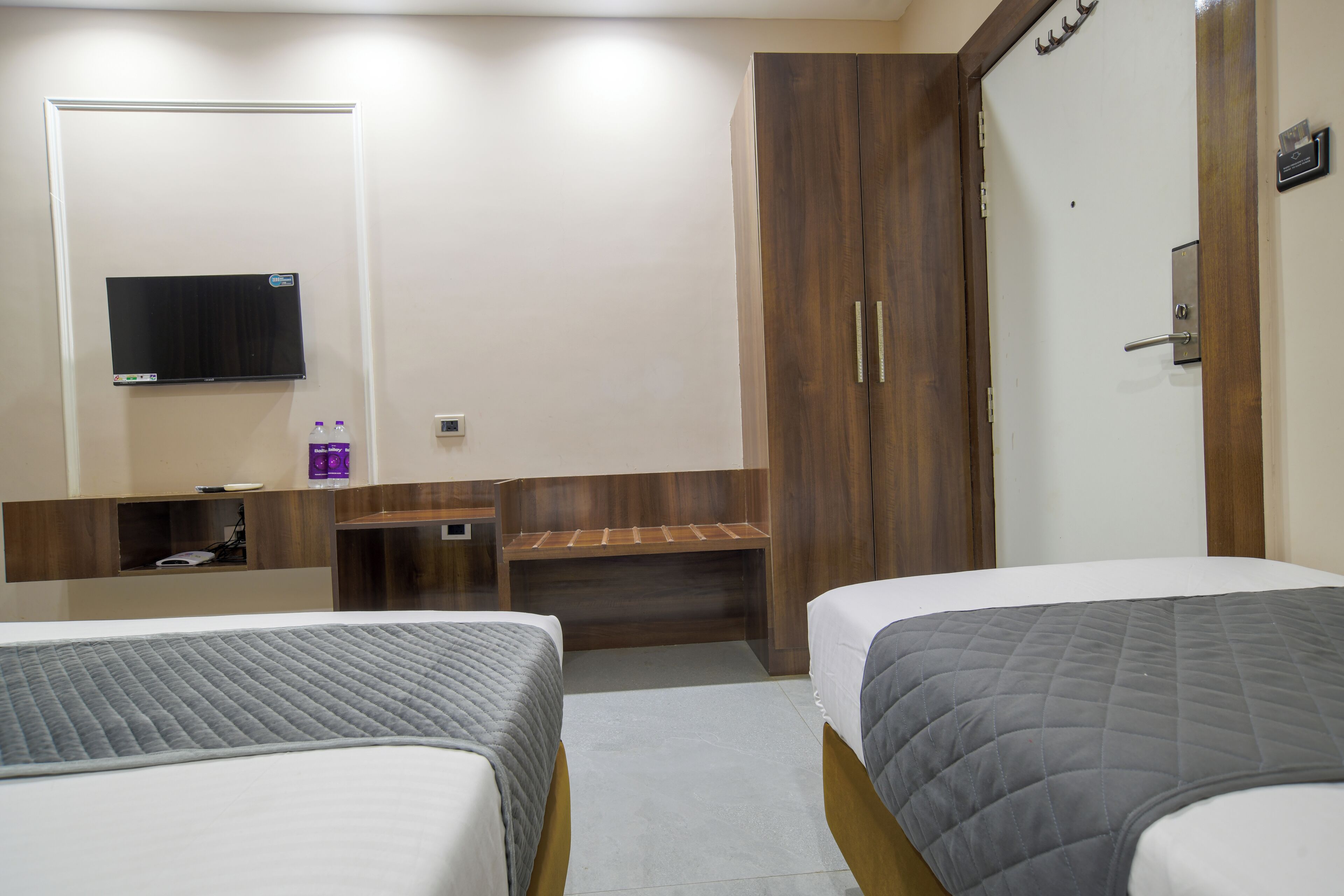 A hotel room with two beds, a TV mounted on the wall, a wooden wardrobe, and a small desk with a chair.