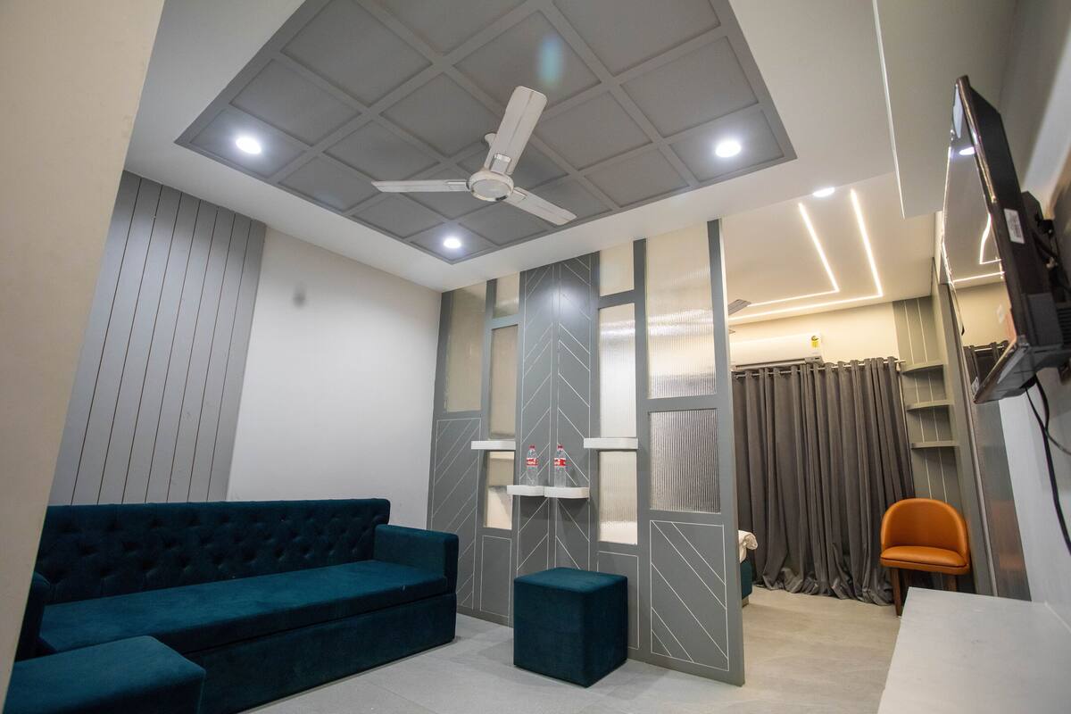 A modern living room with a teal sofa, a small ottoman, a ceiling fan, and a TV mounted on the wall.