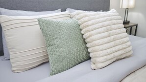 A bed with a grey headboard, a white pillow, a green textured pillow, and a white fluffy blanket. - Molivu Homes 1BR | New Build | Pet Friendly | W/D (San Jose)