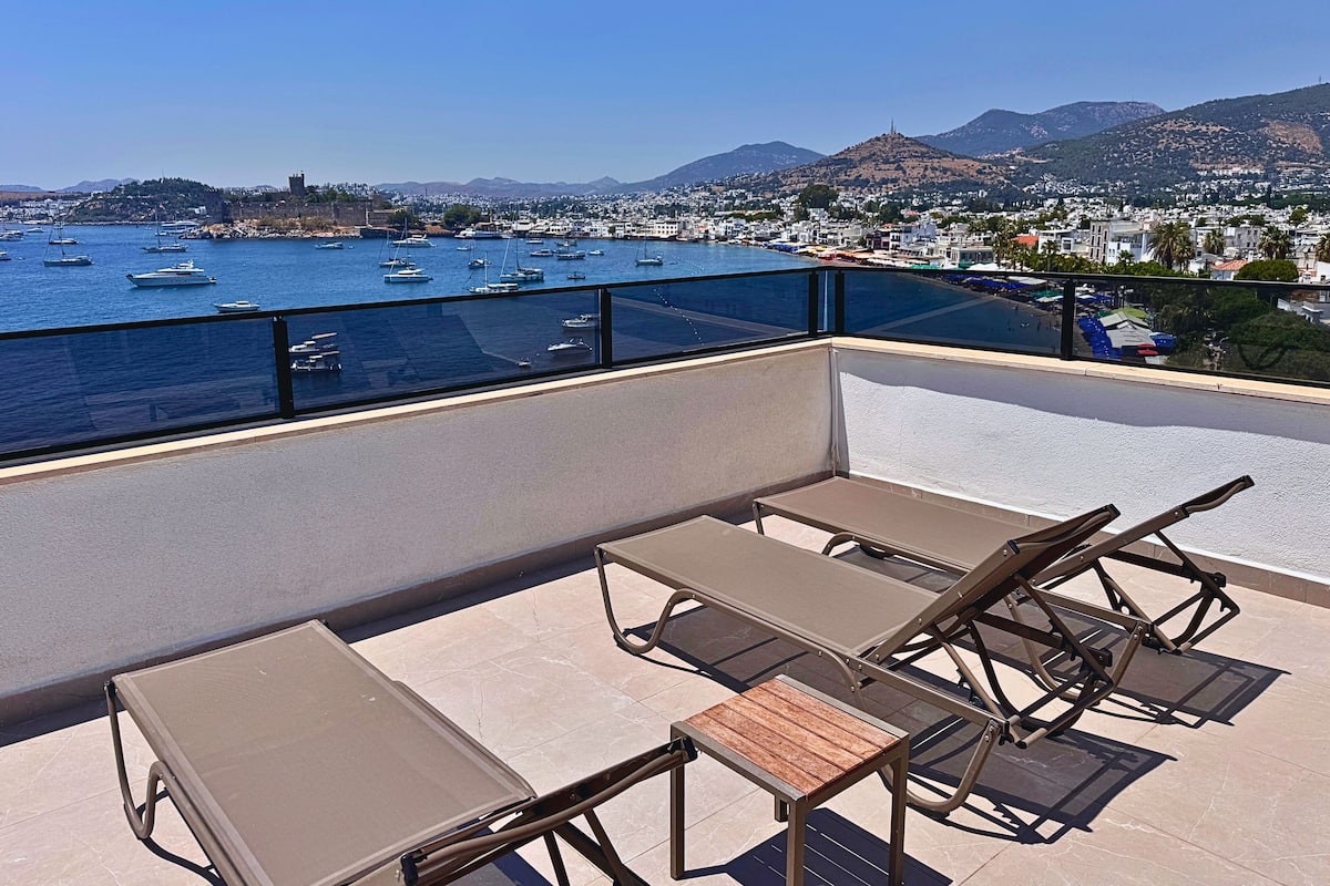 A balcony with two lounge chairs and a small table, overlooking a marina with boats and a coastal town.