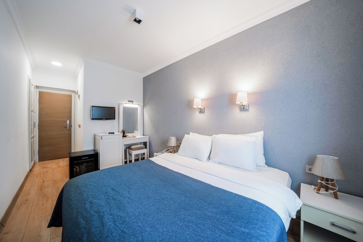 A hotel room with a bed, bedside table, lamp, TV, and a desk with a chair.