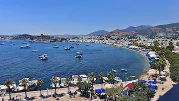 Un pueblo costero con un puerto deportivo, cafés junto a la playa y una vista de la ciudad al fondo.