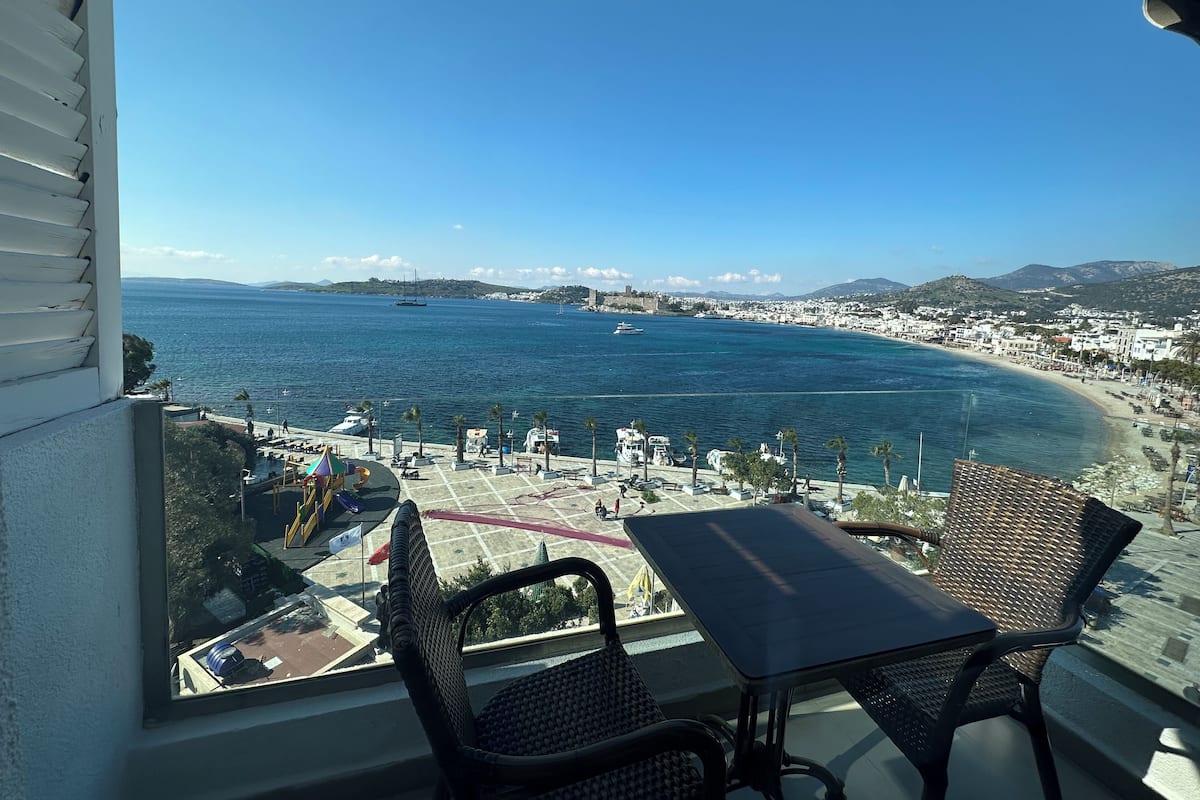 A balcony with wicker chairs and a table overlooking a coastal town with a clear view of the sea and boats.