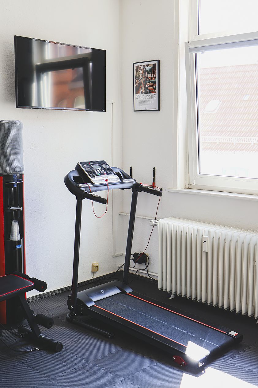 Ein Zimmer mit Laufband, Wandfernseher, Heizkörper und Fenster mit Blick auf Gebäude.