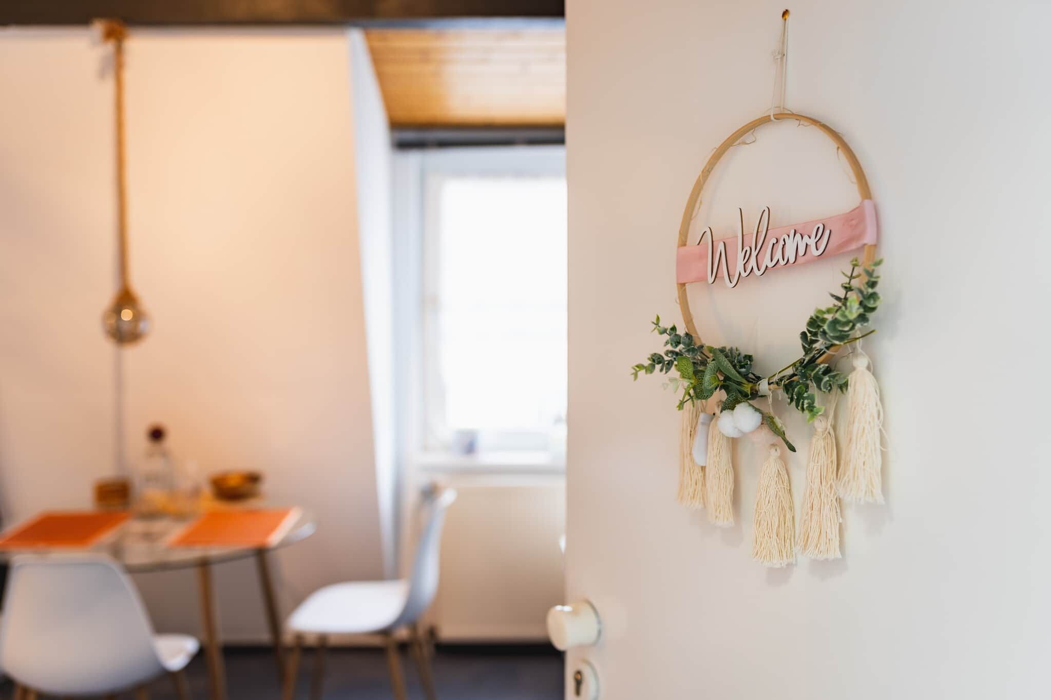 Een kamer met een eettafel, stoelen, een hanglamp en een wanddecoratie met een 'Welcome'-bord en planten.