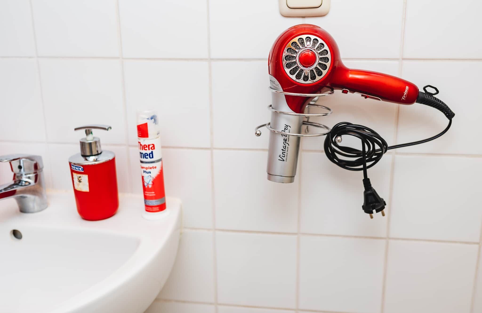 A bathroom with a red hair dryer mounted on the wall, a sink, and two bottles of hand soap.