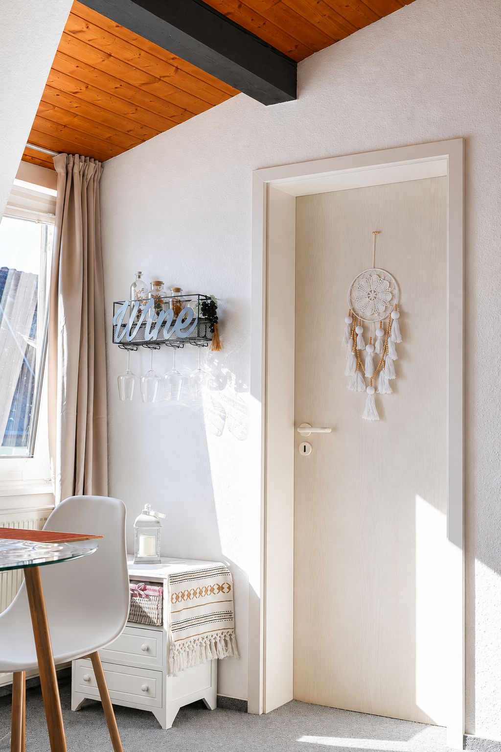 Une pièce avec un plafond en bois, une porte blanche ornée d’un attrape-rêves, un porte-bouteilles fixé au mur et une chaise blanche.
