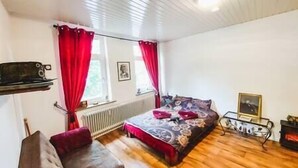 Ett sovrum med en säng, en soffa, ett element, ett fönster med gardiner och en vägghylla.