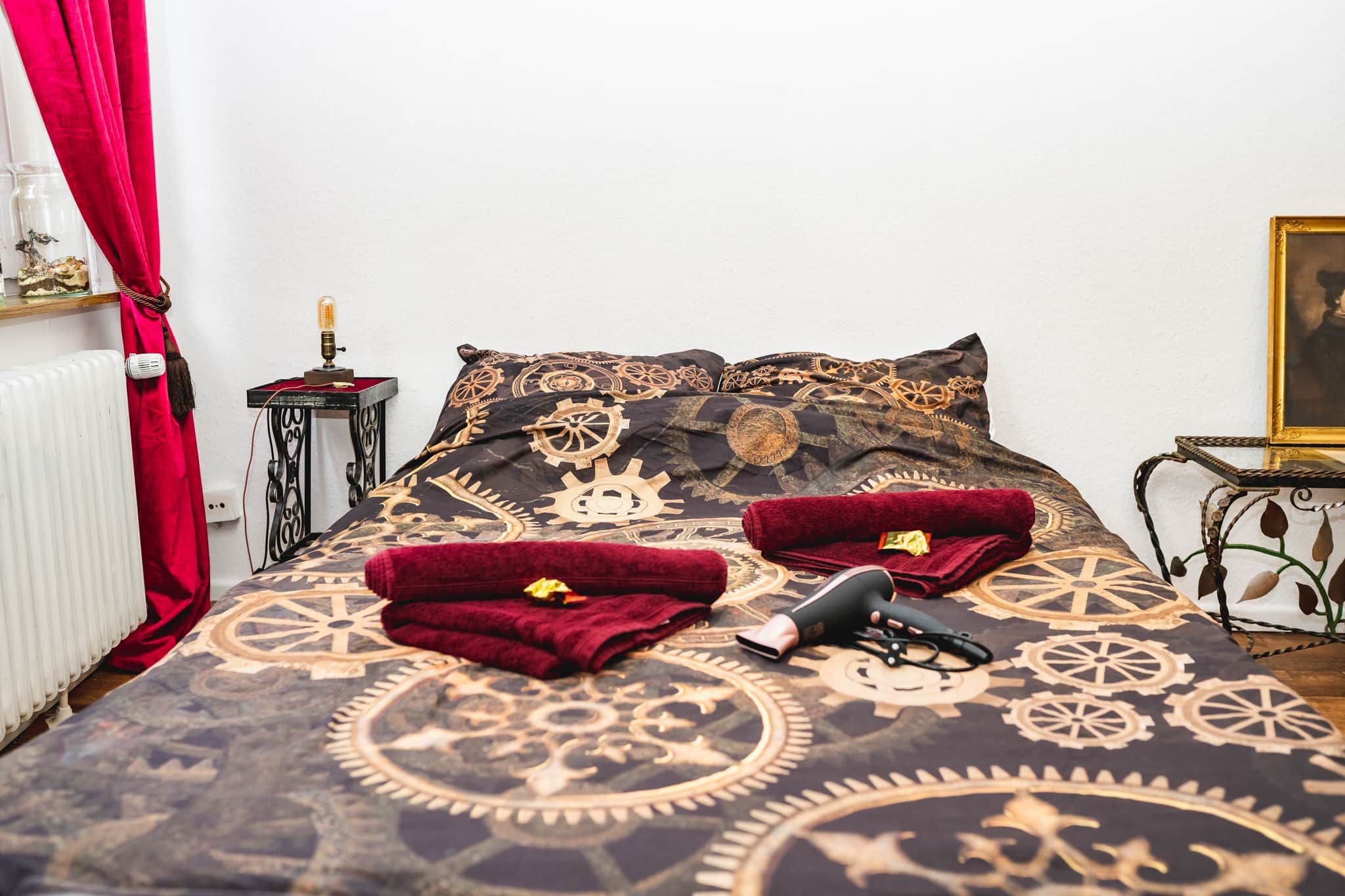 A bed with a patterned bedspread, two red towels, a hairdryer, and a small table with a lamp.