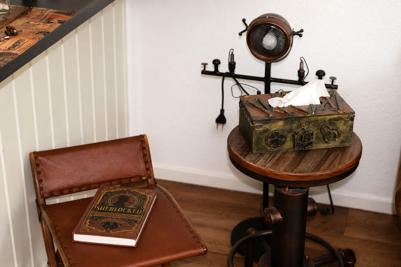 Una scrivania in stile vintage con una poltrona in pelle, un libro intitolato 'Sherlock Holmes' e una lampada da tavolo antica.