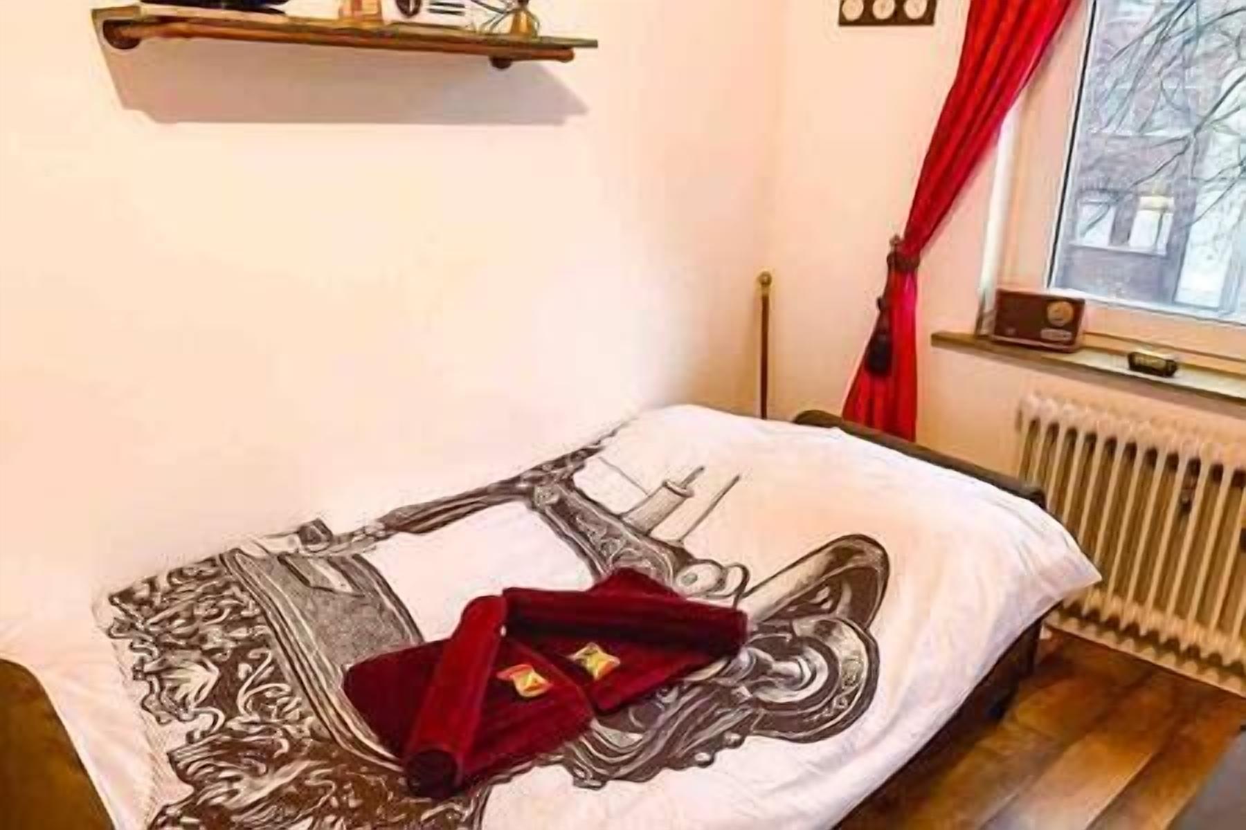 Habitación de hotel con cama que presenta un diseño detallado, una manta roja, un radiador y una ventana con cortinas.