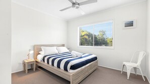 Ein Schlafzimmer mit einem Bett, einem Nachttisch, einem Sessel, einem Deckenventilator, einem Fenster mit Jalousien und einem gerahmten Bild an der Wand.