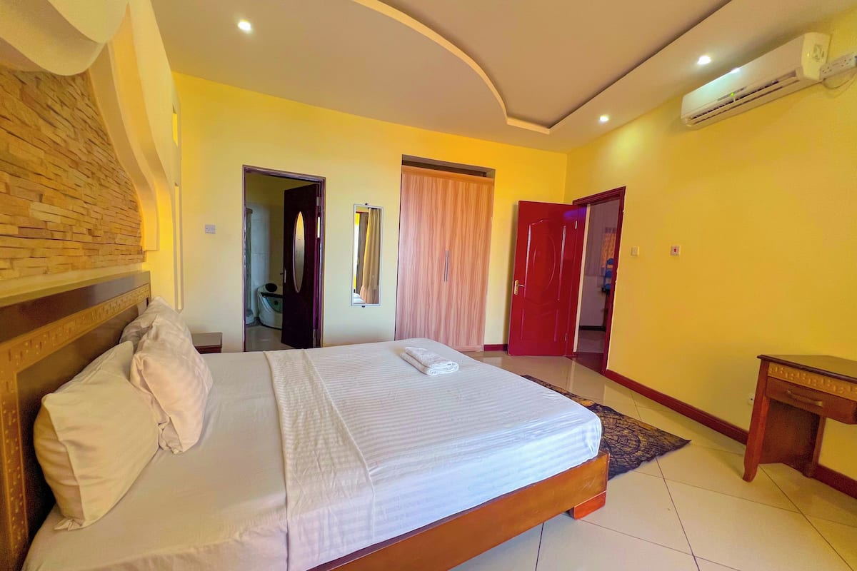 A hotel room with a large bed, a wooden headboard, a mirror, a wardrobe, and an air conditioning unit.