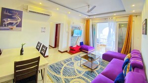 A modern living room with a dining area, purple sofas, a coffee table, and a TV mounted on the wall. - Shanzu Beachfront Apartments (Mombasa)