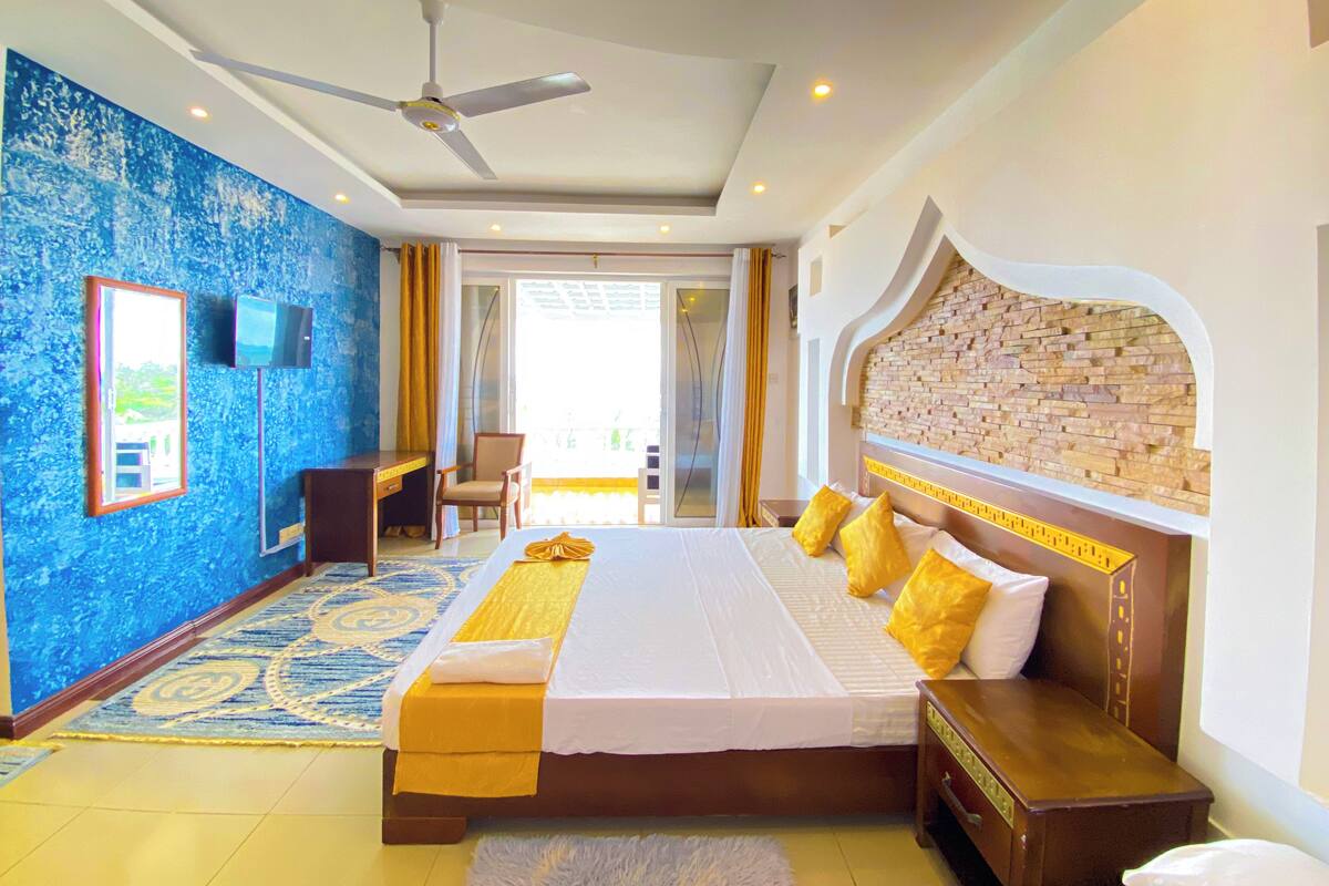 A hotel room with a large bed, a desk, a chair, a TV, and a ceiling fan.