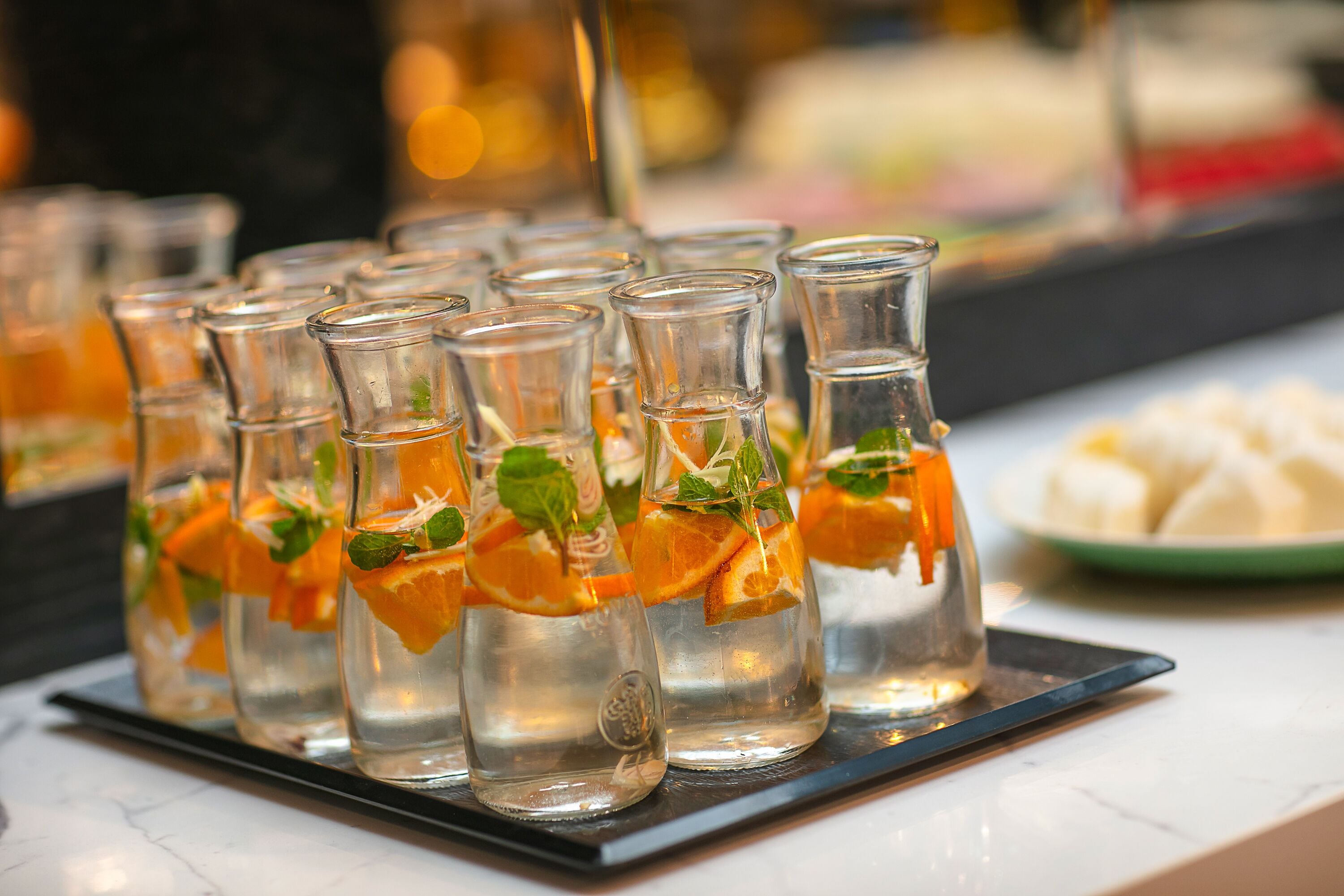A row of clear glass bottles with orange slices and mint leaves, placed on a black tray.