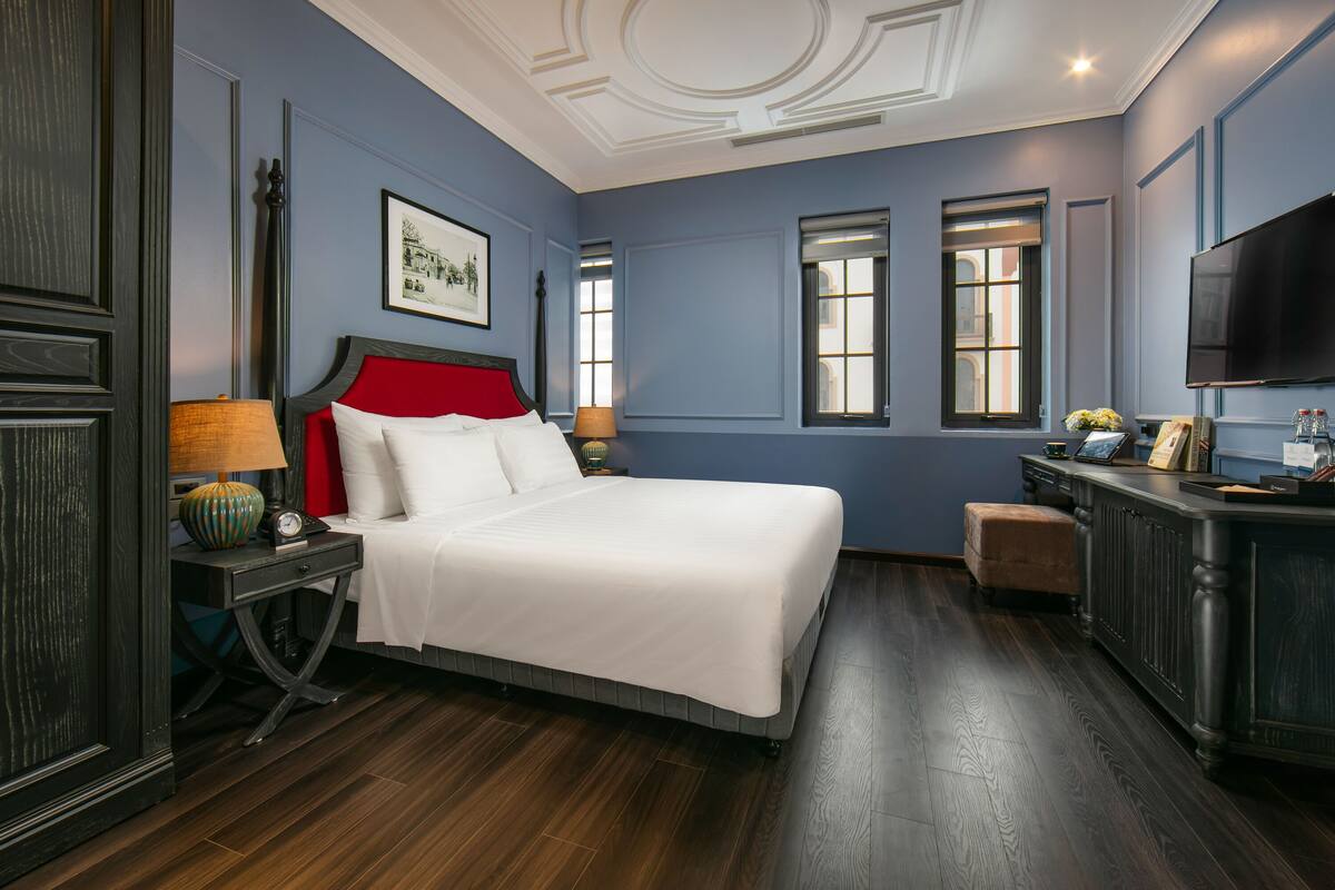 A bedroom with a large bed, a nightstand with a lamp, a dresser, and a television on the wall.
