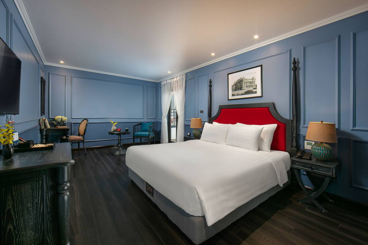 A hotel room with a large bed, a desk, a chair, a small table, and a lamp.