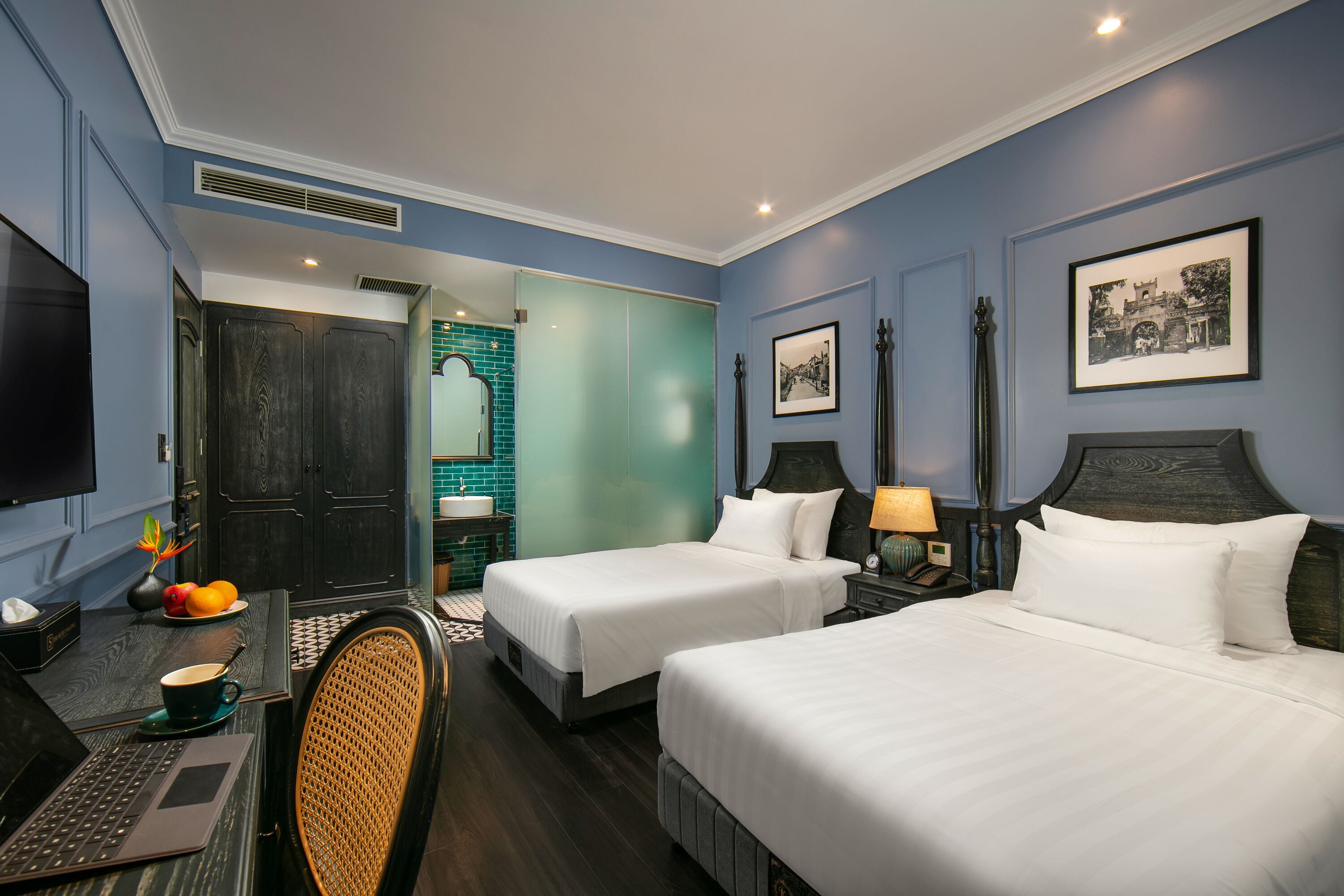 A hotel room with two beds, a desk with a laptop, a chair, a TV, and a bathroom visible through an open door.