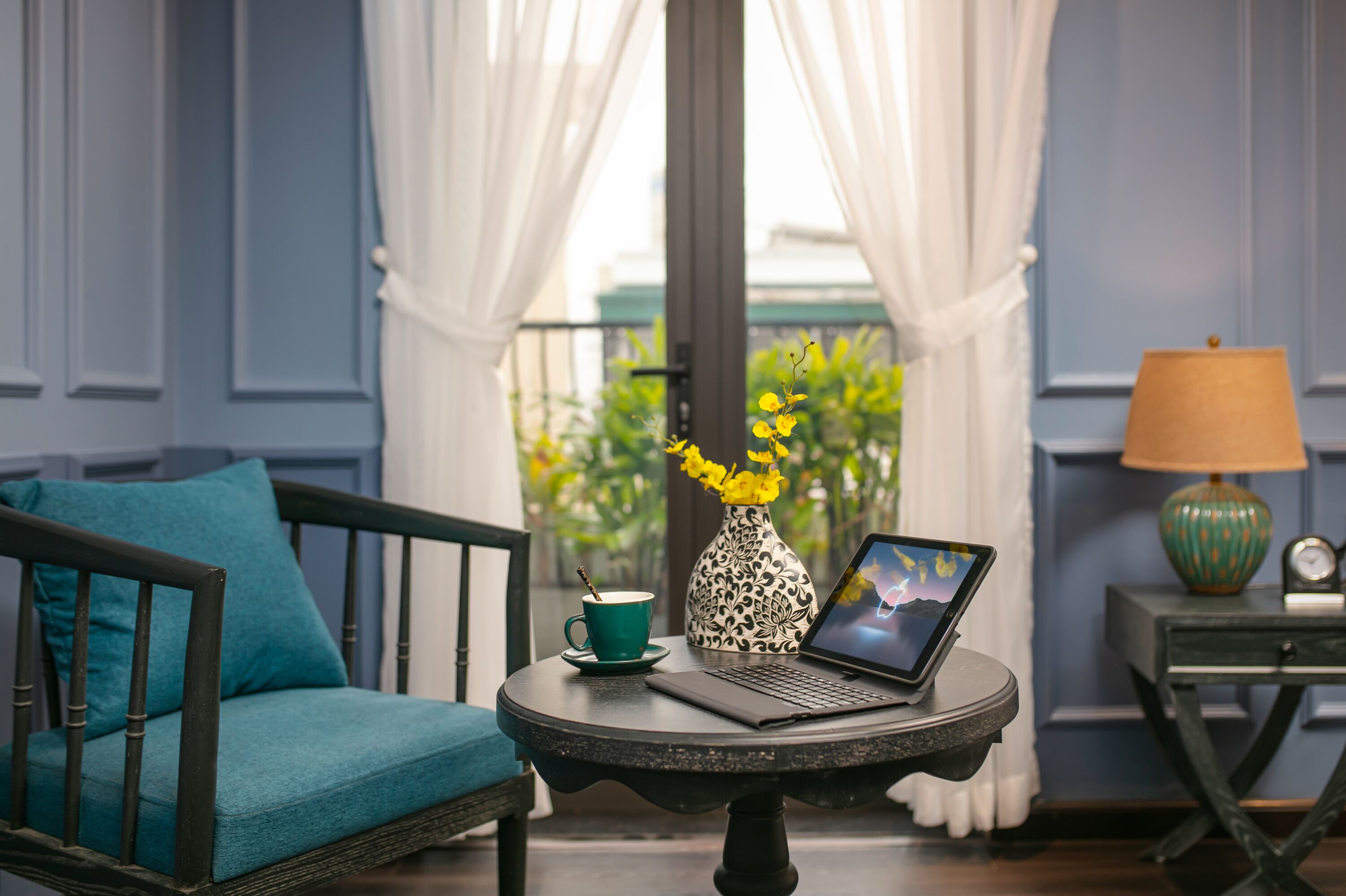 Ein Zimmer mit einem petrolfarbenen Sofa, einem kleinen runden Tisch mit einem Laptop, einer Vase mit gelben Blumen, einer grünen Tasse und einer Lampe.