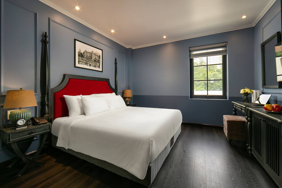 A bedroom with a large bed, a nightstand with a lamp, a dresser, and a window with a view.