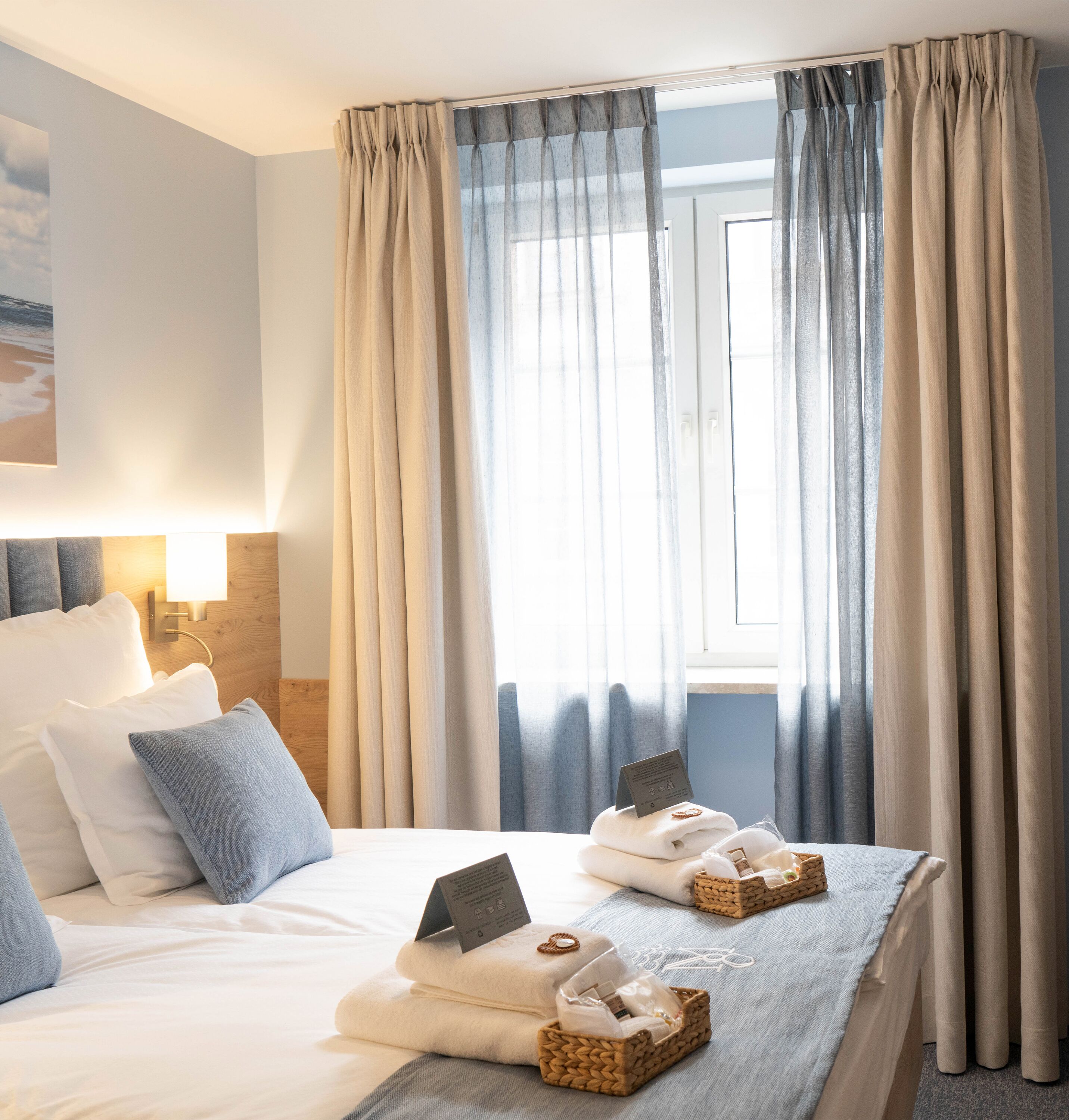 A hotel room with a bed, pillows, a bedside lamp, a window with curtains, and towels in a basket.