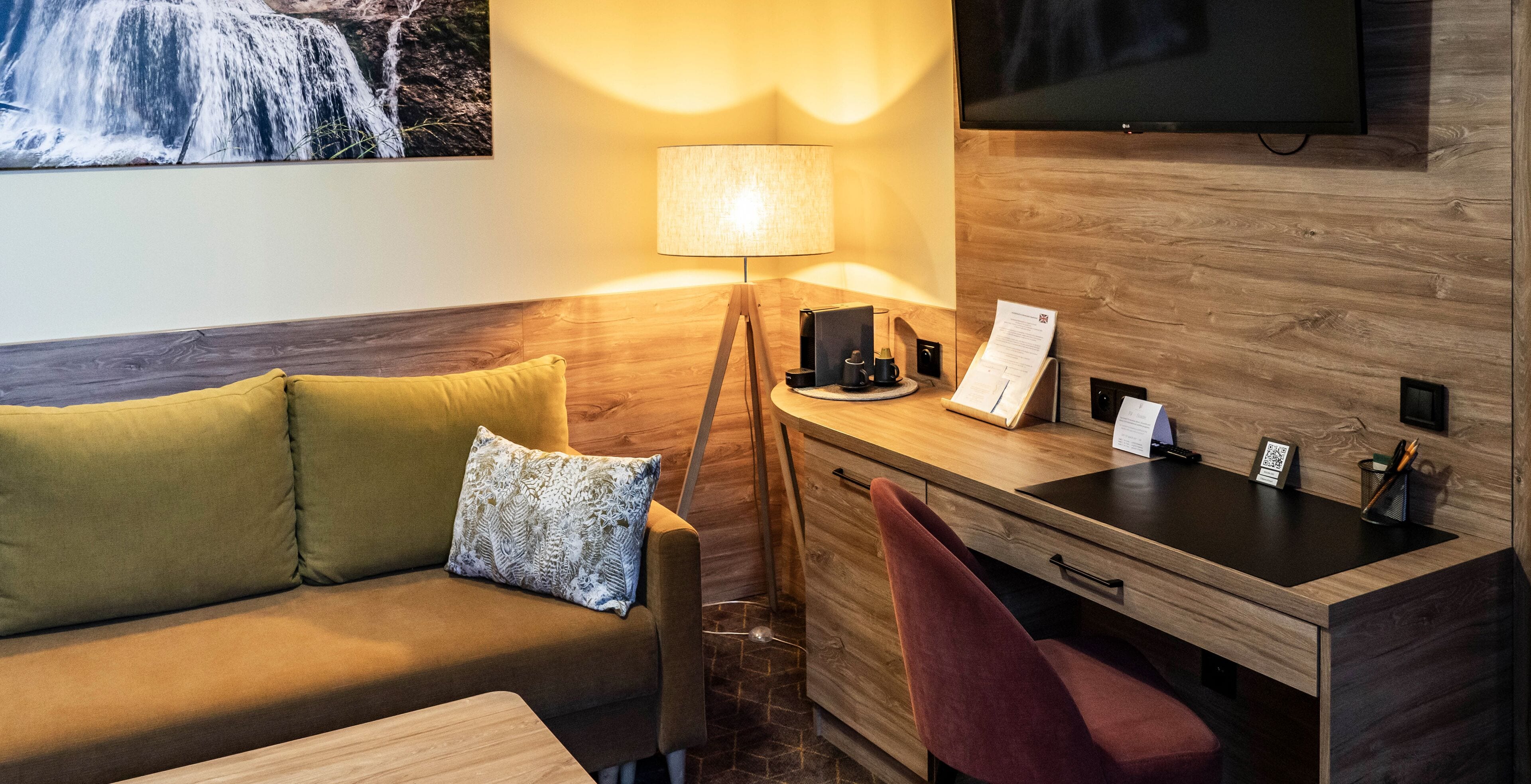 Habitación de hotel con escritorio de madera, sofá con cojines, lámpara, televisor y una obra de arte en la pared.
