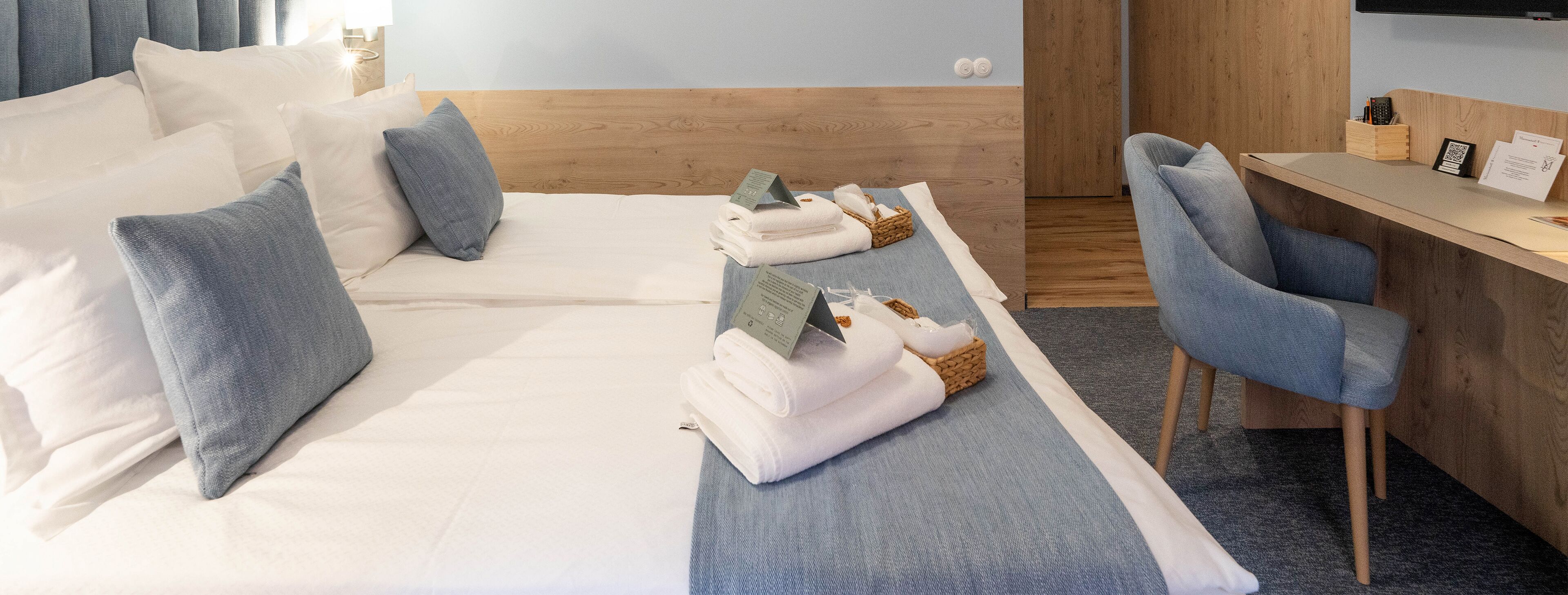 A hotel room with a bed, a desk, and a chair. There are towels and a basket on the bed.