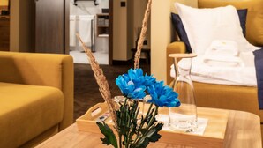 Een hotelkamer met een gele bank, een houten bijzettafeltje met een vaas blauwe bloemen, en een bed met een blauwe deken.