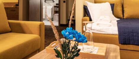 Una habitación de hotel con un sofá amarillo, una mesita de madera con un jarrón de flores azules y una cama con una manta azul.