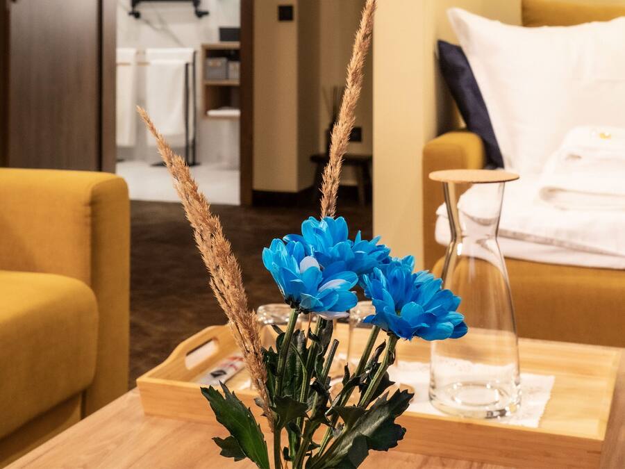 A hotel room with a yellow sofa, a wooden side table with a vase of blue flowers, and a bed with a blue blanket.