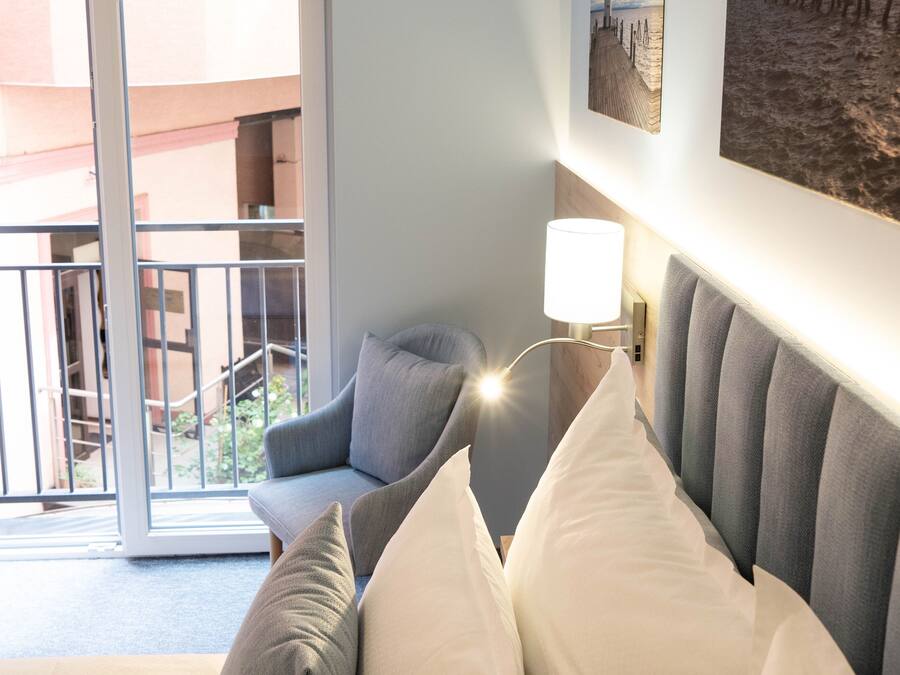 A hotel room with a bed, a chair, a wall-mounted lamp, and a view of the outside balcony.