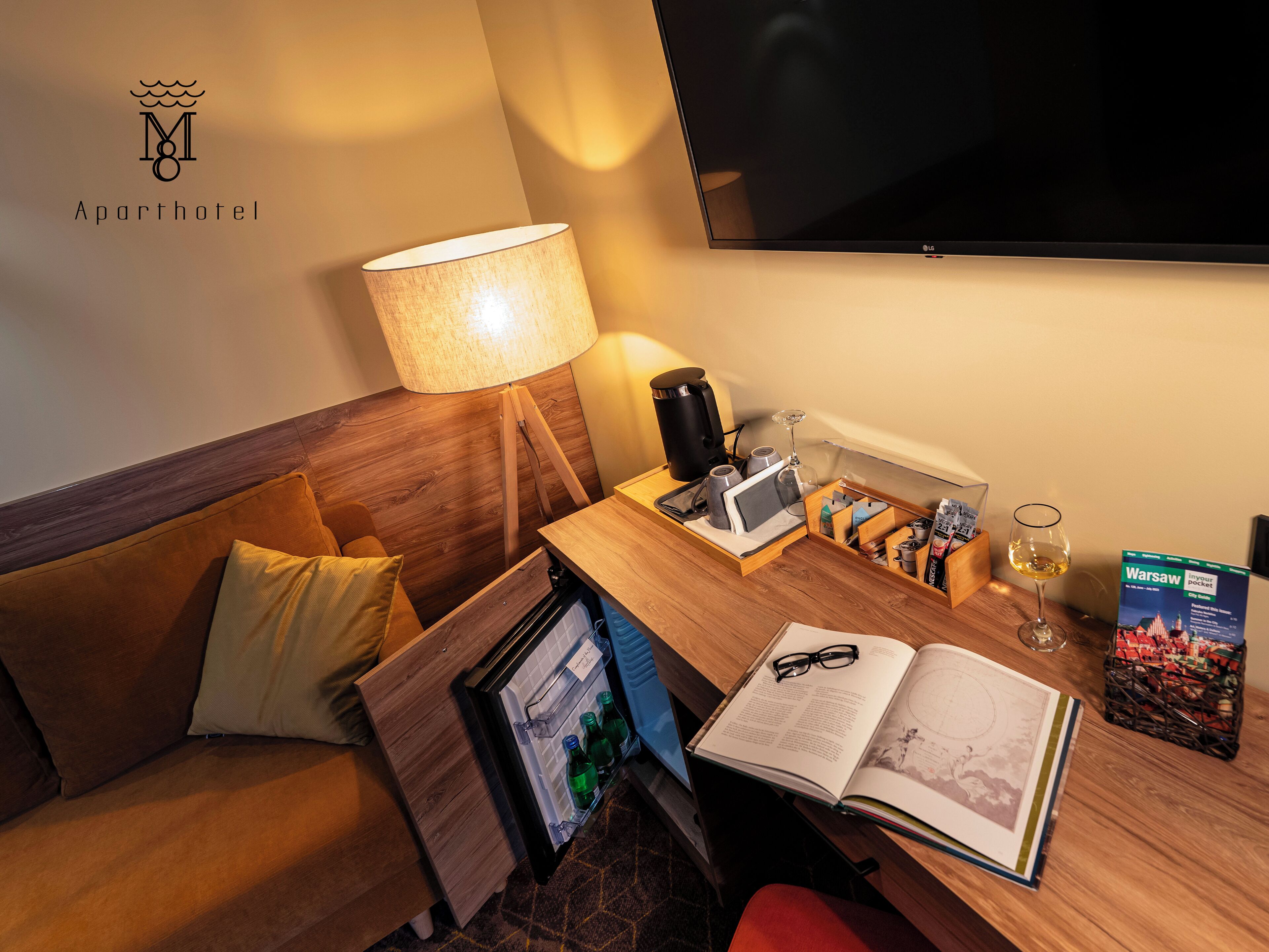 Habitació d'hotel amb un capçal de llit de fusta, un sofà, una taula de treball amb una làmpada, una cafetera, un got de vi, un llibre i un televisor.