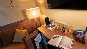 Habitació d'hotel amb un capçal de llit de fusta, un sofà, una taula de treball amb una làmpada, una cafetera, un got de vi, un llibre i un televisor.