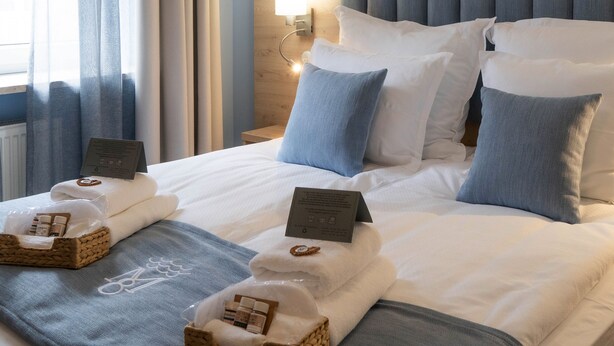 Een net opgemaakt bed met witte lakens, blauwe kussens en een nachtkastje met een lamp.