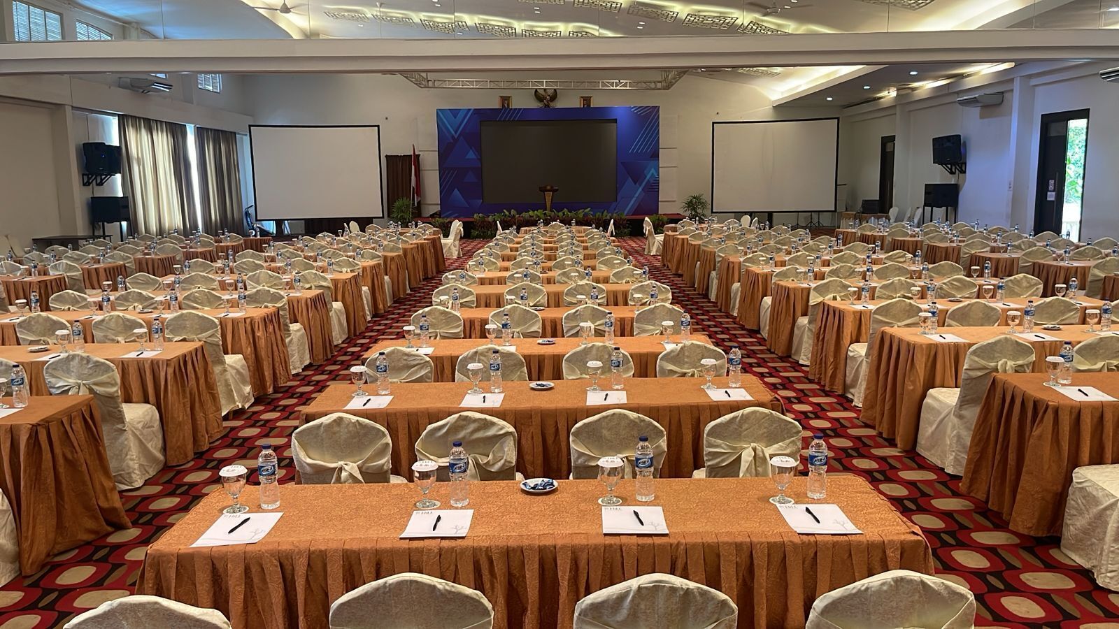 A large conference room with rows of tables and chairs, equipped with water bottles and name cards.