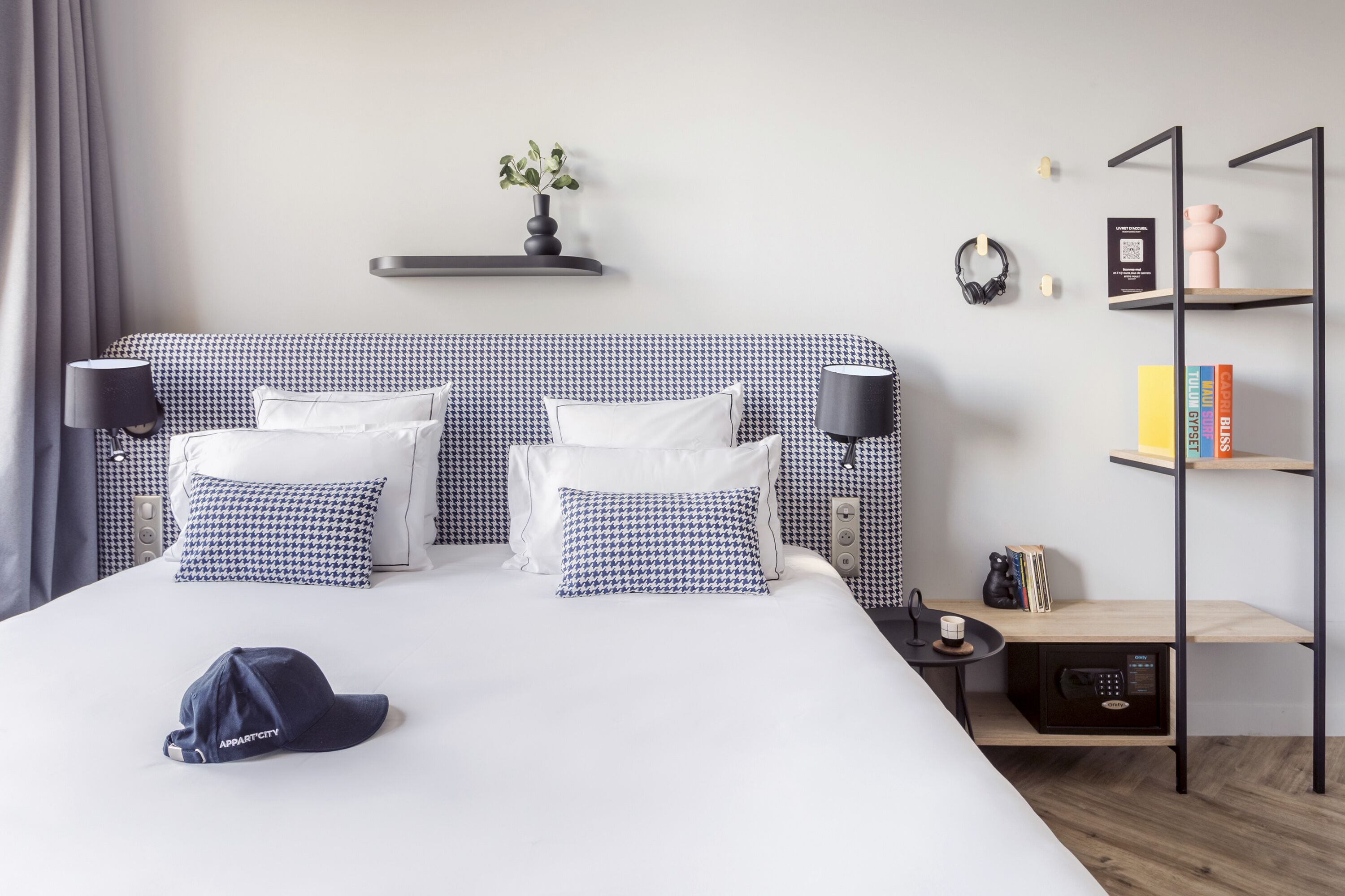 Ett hotellrum med en stor säng, två sänglampor, ett litet bord med en kaffebryggare, en bokhylla och en vägghylla med dekorativa föremål.