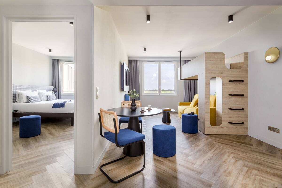Habitació d'hotel moderna amb terra de fusta, taula de dinar rodona, cadires blava tapissades, un sofà i un moble de fusta únic amb calaixos.