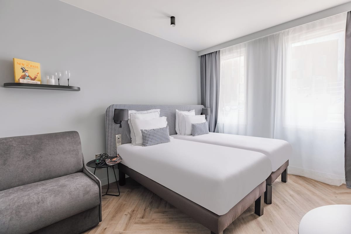 Habitació d'hotel moderna amb un llit gran, un sofà gris, una taula petita amb una làmpada i una estant amb llibres i vasos.